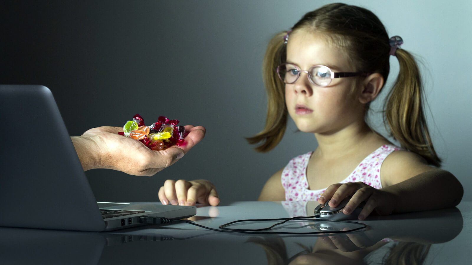 menina frente ao computador e uma mão oferecendo doces atrativos a ela oferece perigo - O Excesso de Telas na Infância é Um Perigo Silencioso