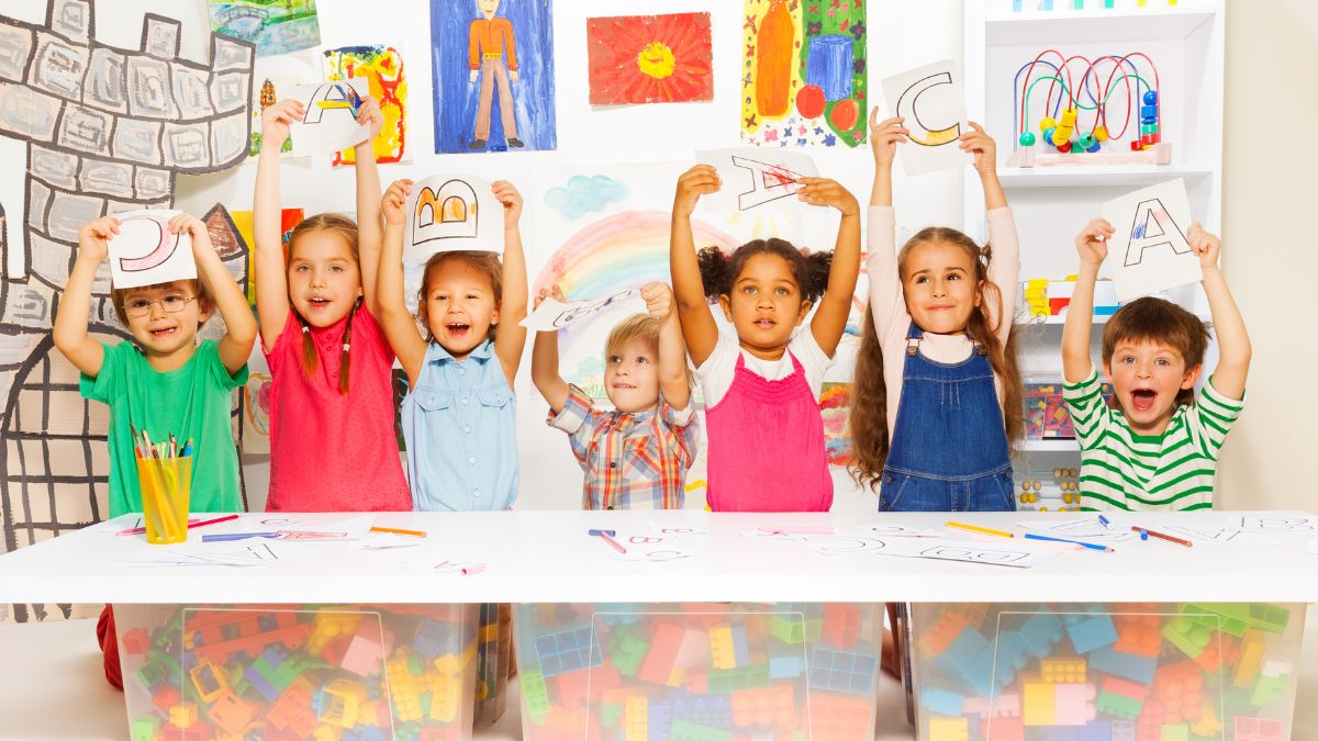 Crianças com braços erguidos e com papel na mão mostrando as letras do alfabeto.
Imagem: Sergey Novikov